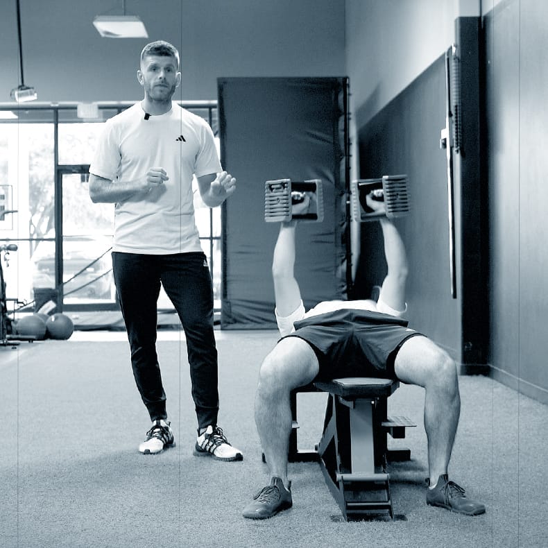 Andy Chadwick is supervising a workout session in a gym. The trainer, dressed in a white shirt and dark pants, is standing next to a person lying on a bench performing a dumbbell chest press. The person on the bench is lifting two large dumbbells above their chest. The gym environment features large windows and exercise equipment in the background. The image is in black and white.