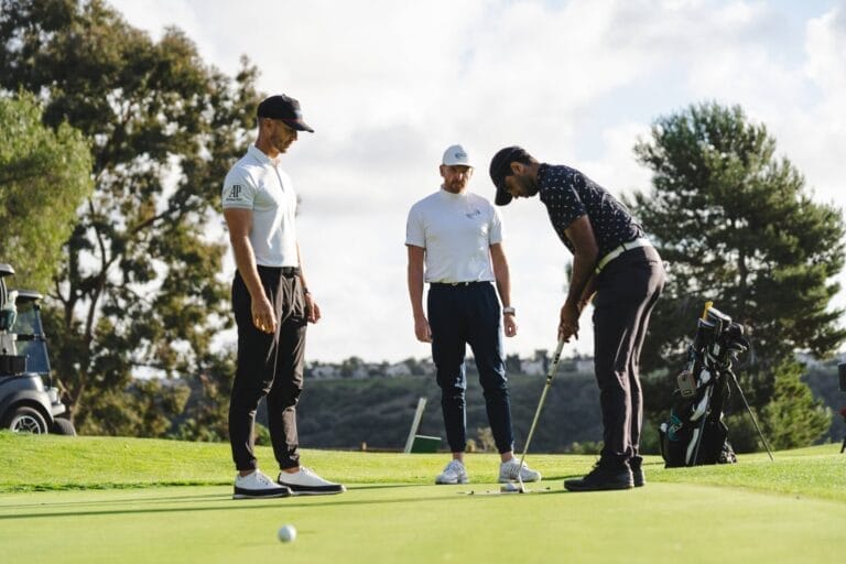 Aaron Rai practices putting on a green, observed by PGA Tour coaches, Piers Ward and Andy Proudman, on a golf course, emphasising precision and technique.