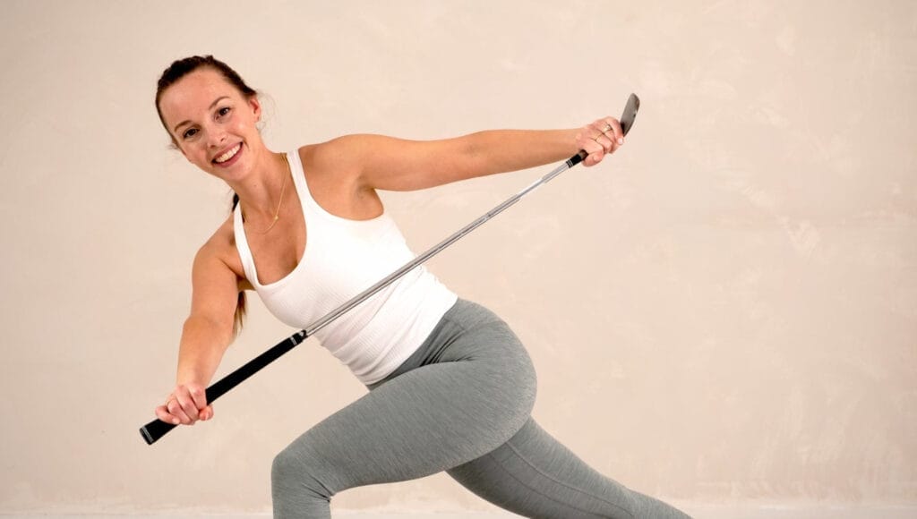 Liesbeth Pauwels is performing a fitness exercise with a golf club. She is smiling, wearing a white tank top and grey leggings. She is in a dynamic pose, lunging with her arms extended, holding the golf club horizontally in front of her. The background is plain and light-colored, emphasizing her movements.