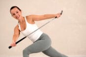 Liesbeth Pauwels is performing a fitness exercise with a golf club. She is smiling, wearing a white tank top and grey leggings. She is in a dynamic pose, lunging with her arms extended, holding the golf club horizontally in front of her. The background is plain and light-colored, emphasizing her movements.