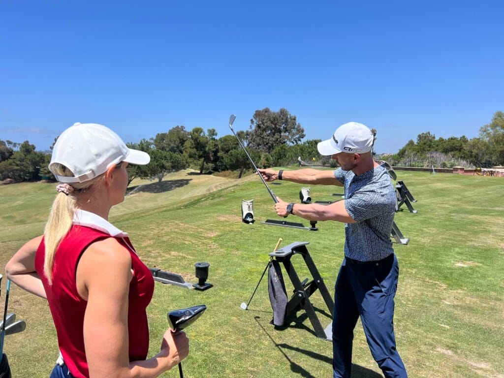 A woman watches as golf coach Andy Proudman demonstrates a swing technique on a sunny driving range.